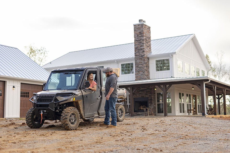 A woman in an ATV talks out the window to a man holding a bag of concrete mix on his shoulder.