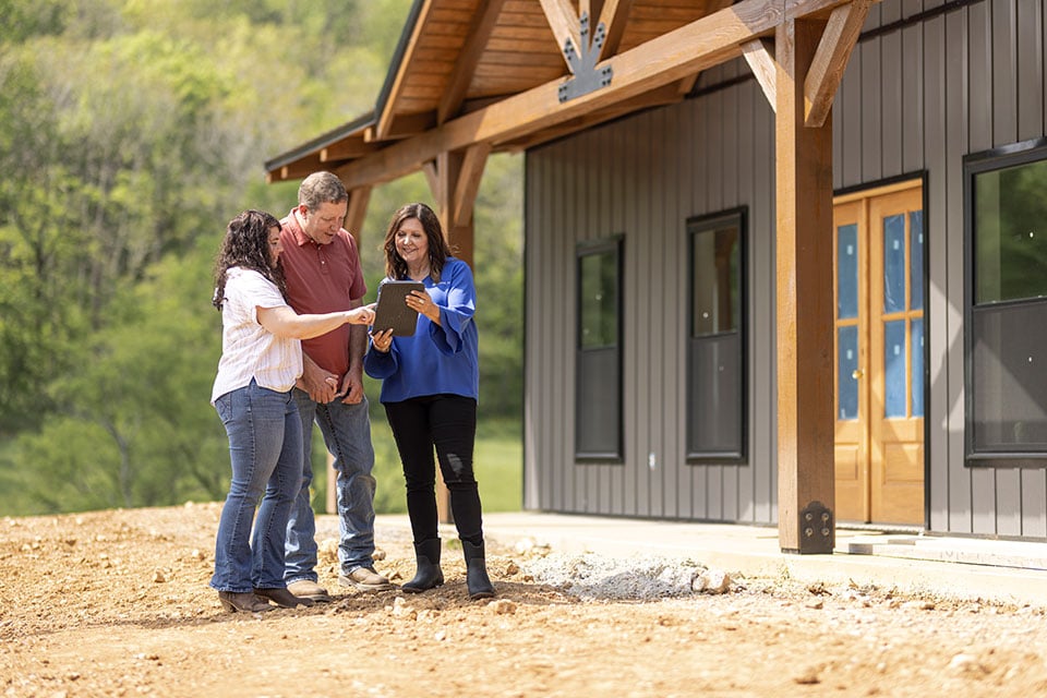 A woman shows something on a tablet screen to a man and woman outside a newly constructed barndominium.