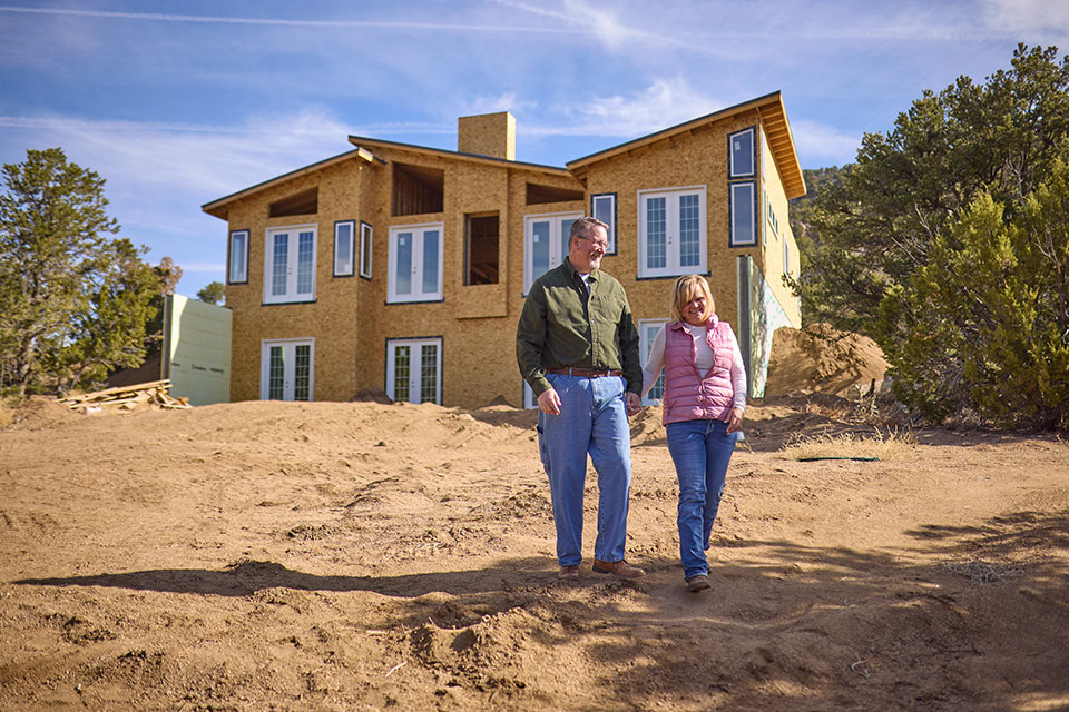 A couple walks across a dirt lot with their unfinished home construction behind them.