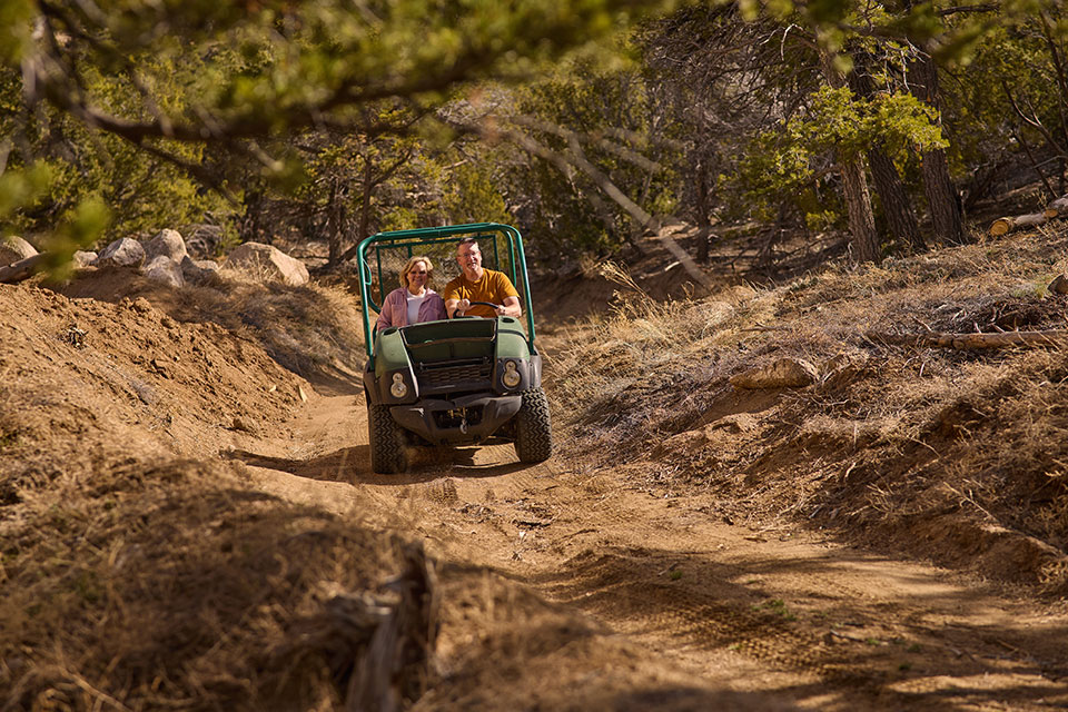 A man and woman drive an ATV along a dirt road in the woods.