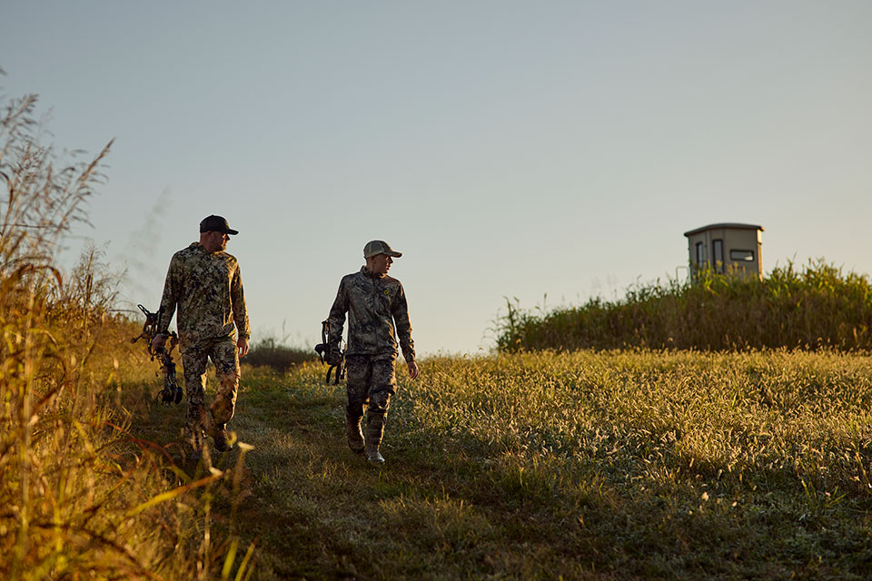 Two men in camo with hunting bows walk casually down a grassy slope at sunset.