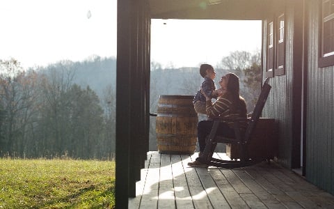 A mom holding her son on the porch of their new rural home.