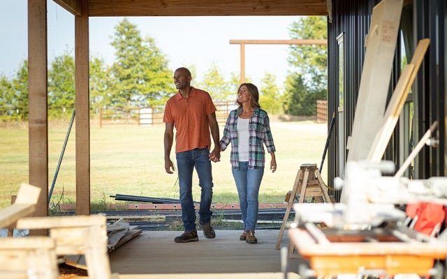 A couple holding hands, walking on the porch of their under-construction rural home.