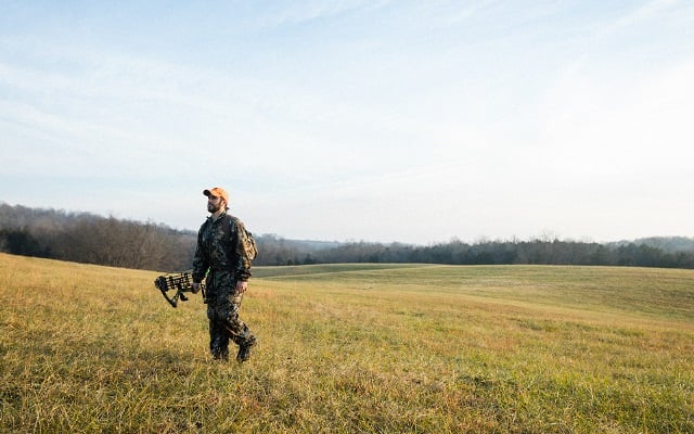 A man dressed in camouflage walking through a rural field carrying hunting equipment.