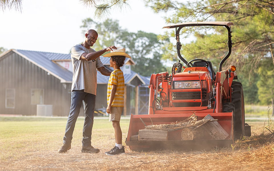 A man putting a hat on his son in front of a tractor with a bucket, with their rural home in the background.