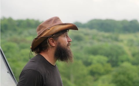 A man, in profile, with a big beard looking off into the distance, with woods beyond him.