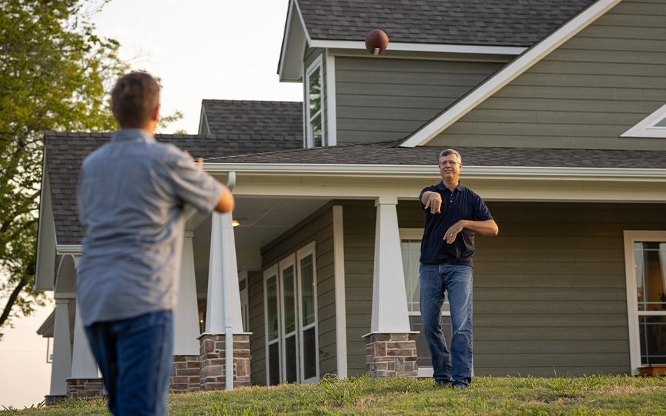 A father and son throwing a football in the yard of their newly built rural home.