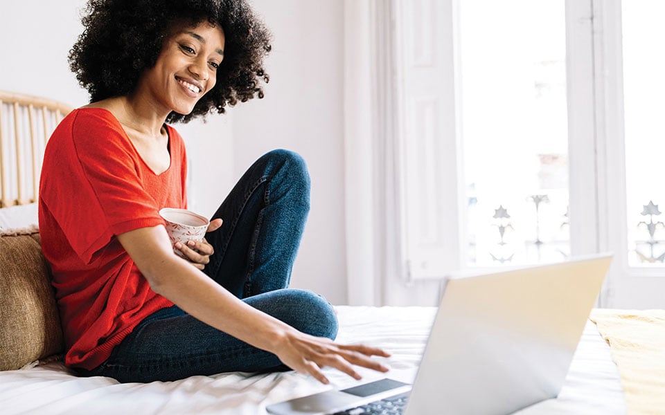 A woman sitting on her bed smiling while using her laptop.