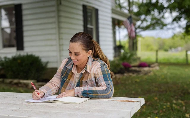 A woman writes in a notebook on a picnic bench outside a home.